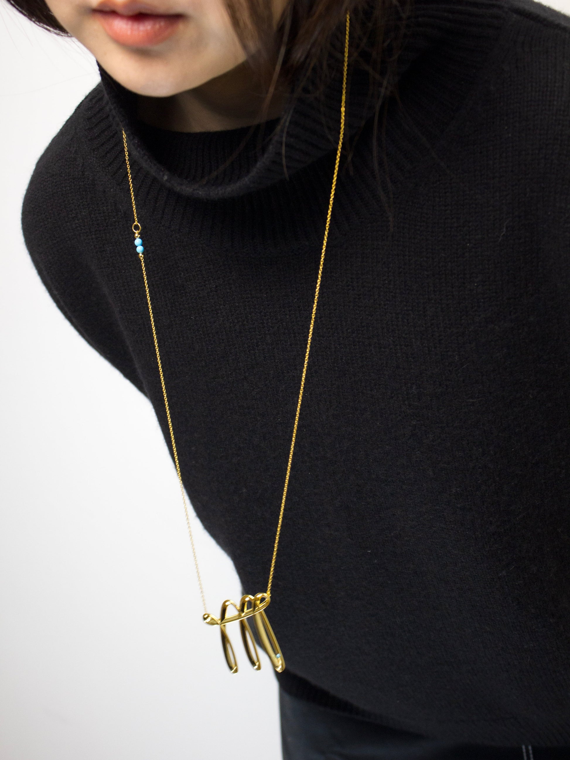 Person wearing a black sweater with a gold necklace featuring a dynamic unique pendant with blue turquoise.