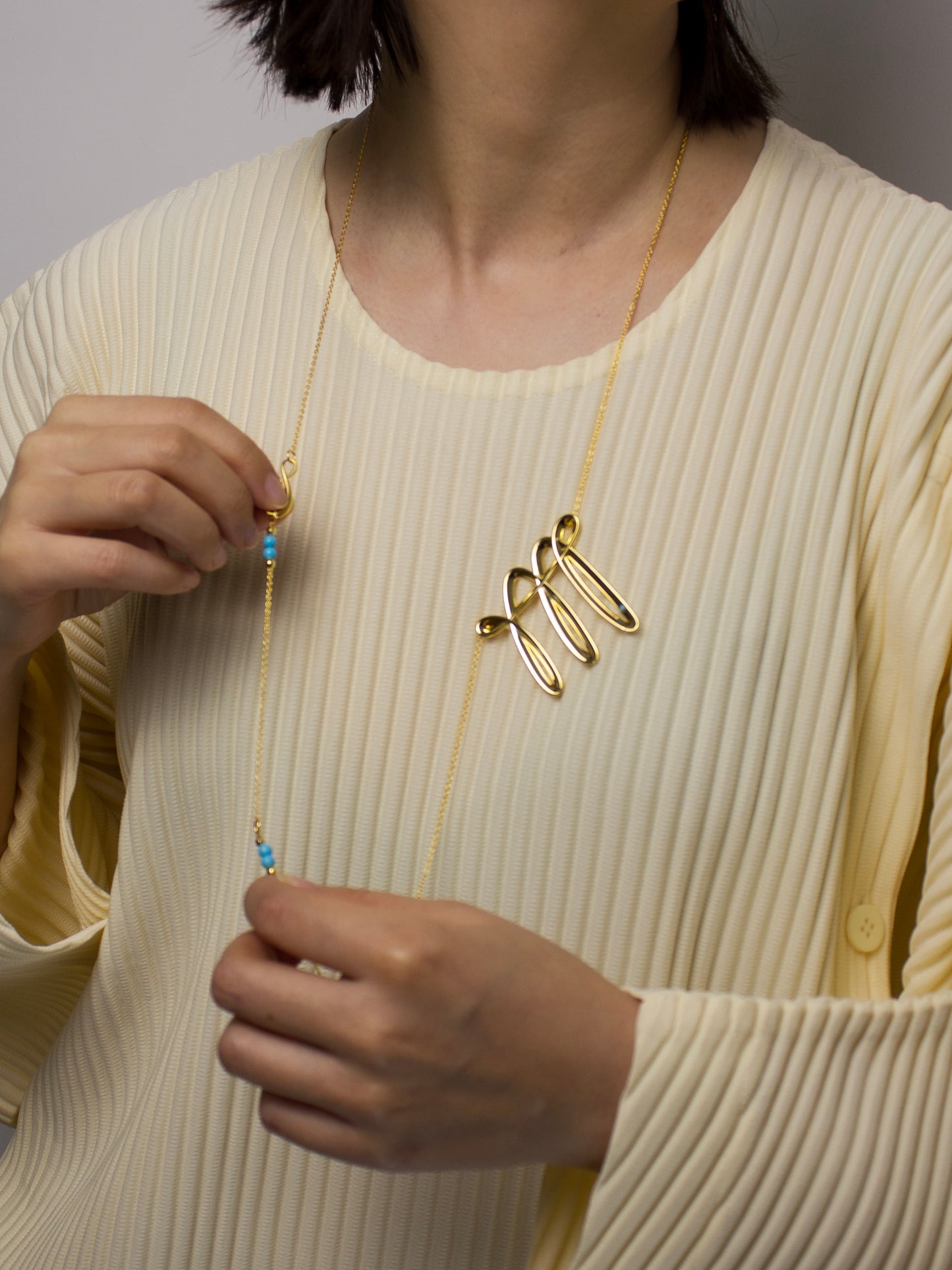 Person wearing a long gold necklace with a woven looking unique design, holding the adjustable chain with turquoise details.