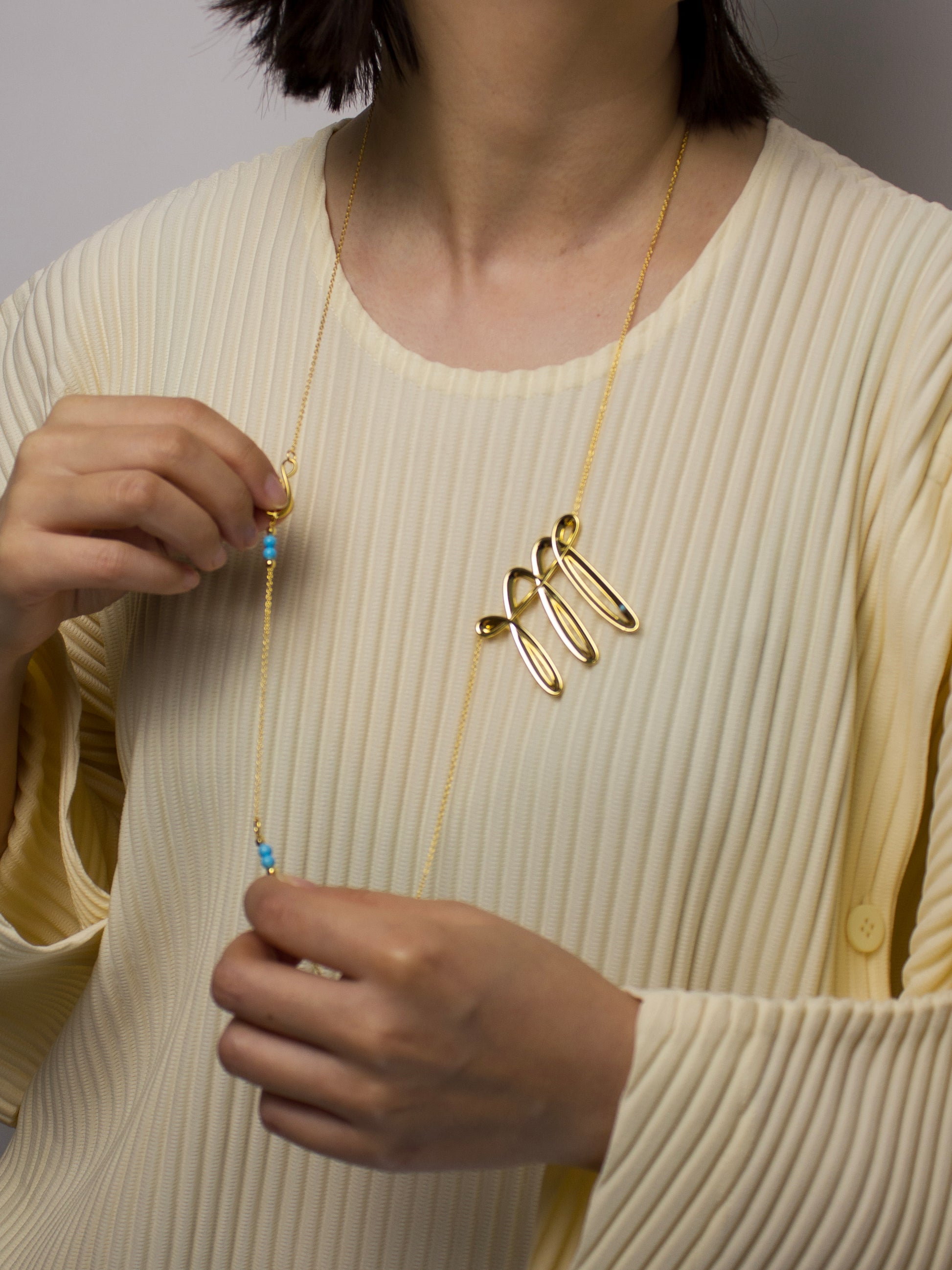 Person wearing a long gold necklace with a woven looking unique design, holding the adjustable chain with turquoise details.